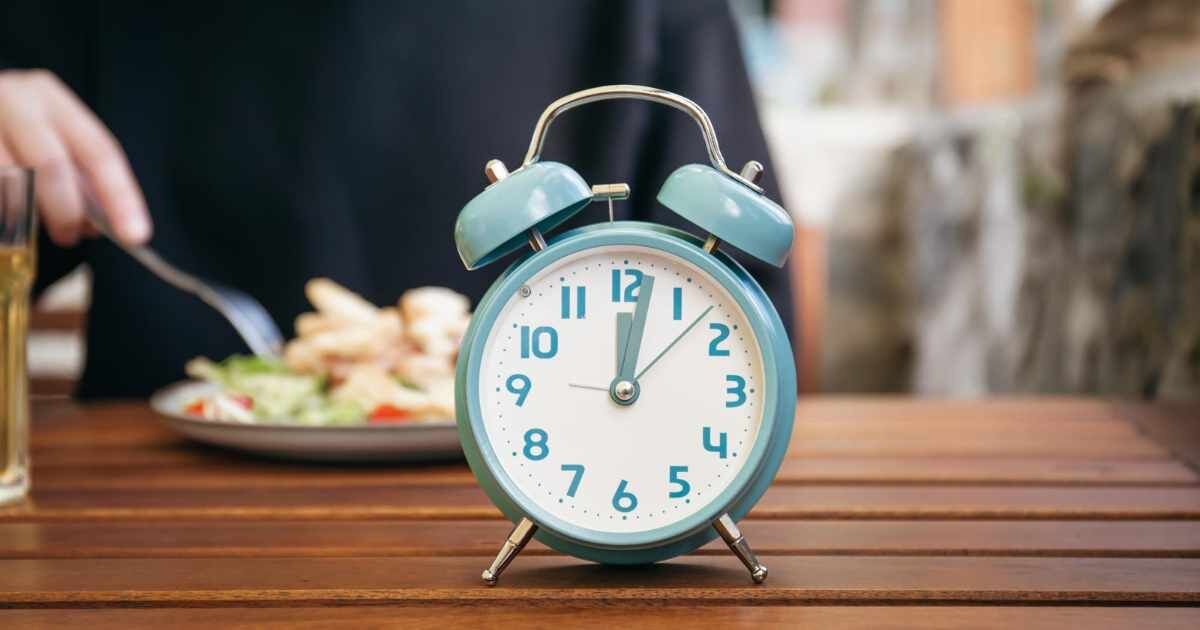 Alarm clock on a wooden table with a plate of food blurred in the background.